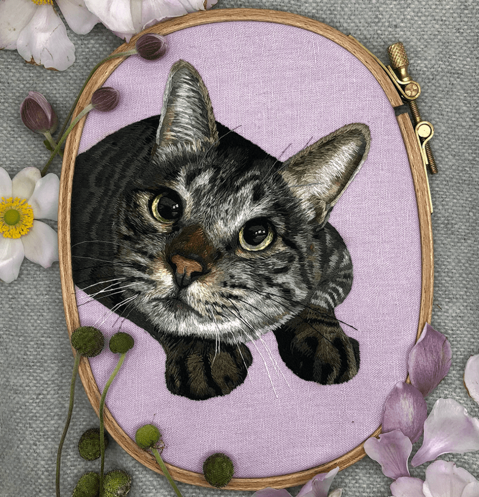 A photo of an embroidered tabby cat. The cat is crouching and has huge, open eyes.